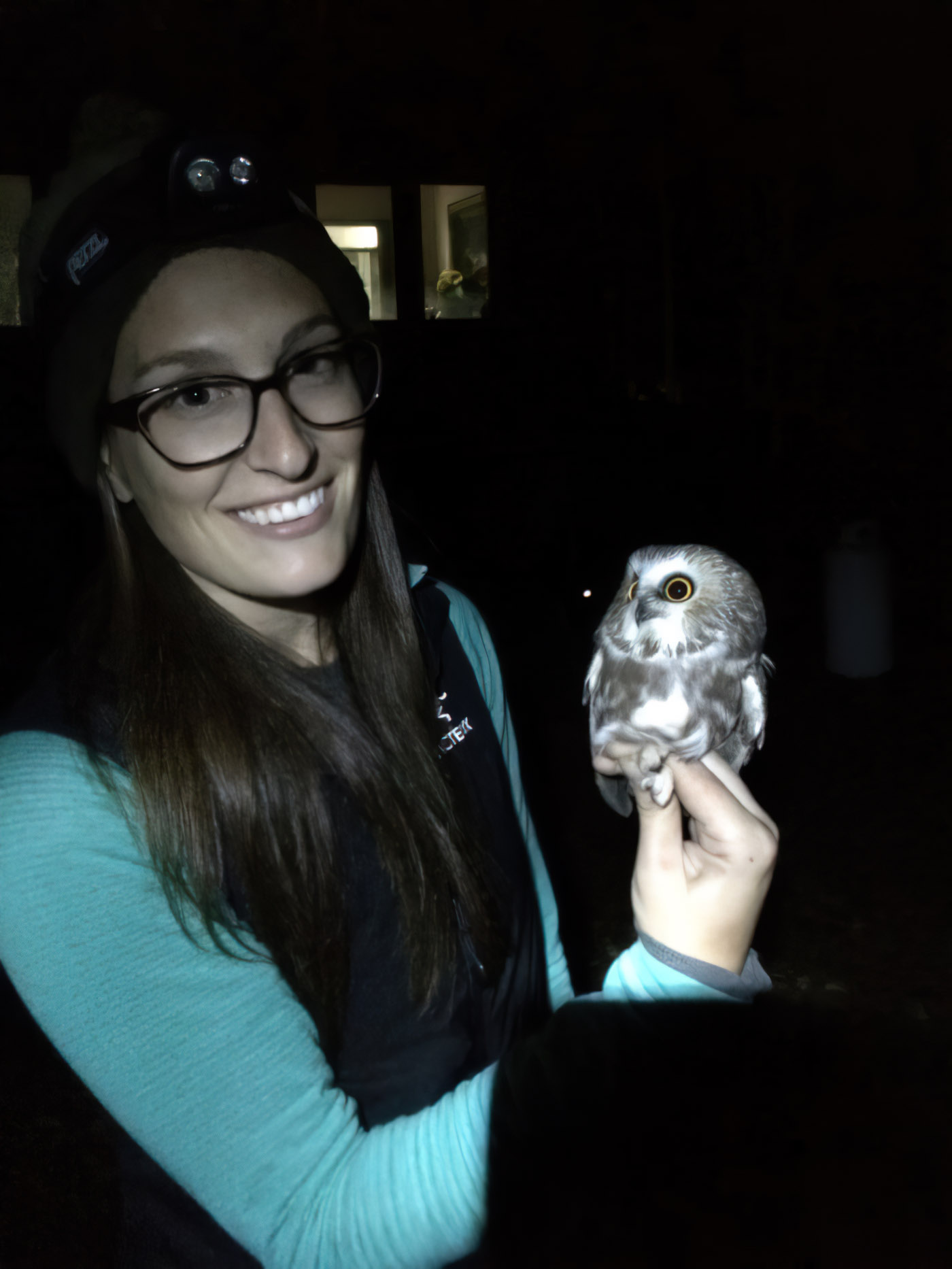 Natasha Crosland, a woman with long brown hair and glasses, wearing a teal shirt and a black vest, in a dark room holding an owl.