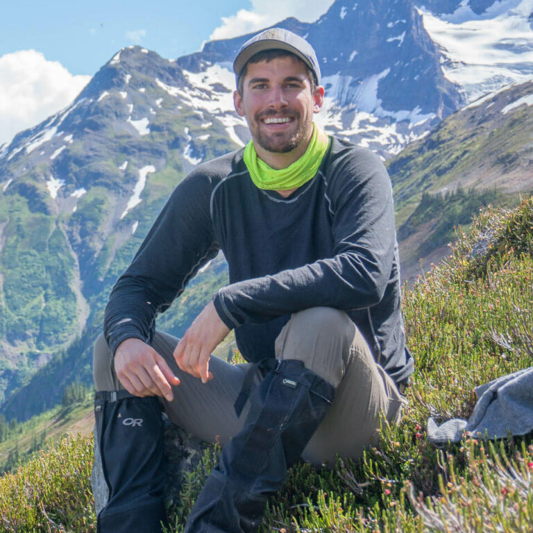 Dr. Clayton Lamb, a man with short hair and stubble, is sitting on a green hillside, in front of snowcapped mountains, and wearing a gray shirt, brown pants, a bright green cowl, and gray baseball hat.