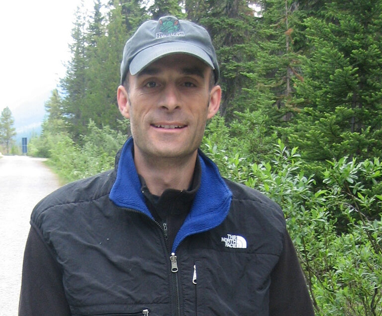 Dr. Craig Demars is standing on a road next to a heavily wooded area wearing a black jacket and gray baseball cap.