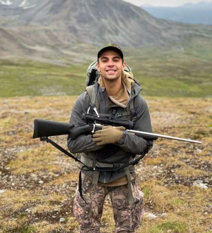 Mateen Hessami is wearing a gray jacket, camo pants, baseball cap, and hiking pack, holding a rifle in an open field at the base of a mountain.